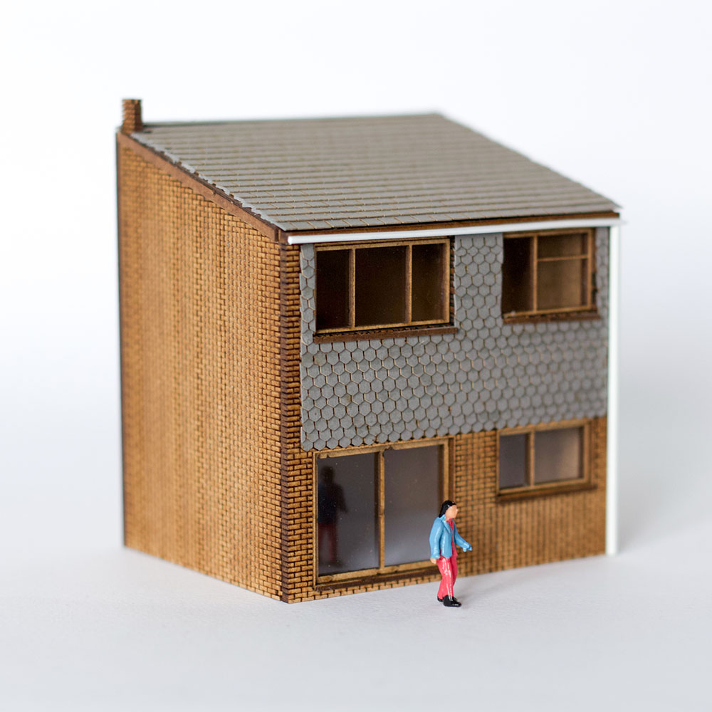 OO gauge relief model of a 1960's mid-century style small house designed by Studio Denley and made from laser-cut MDF by Rail Model. The model is shown at three quarter view and is unfinished showing the cut wood and paper texture of roof and wall tiles. A model figure of a man in a blue jacket and red trousers is stood in front of the model.